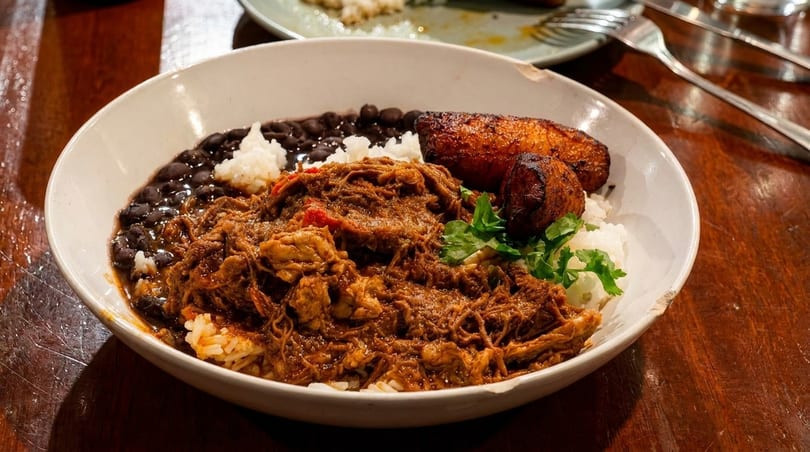 A bowl of ropa vieja with rice, black beans, and fried plantains on a wooden table.