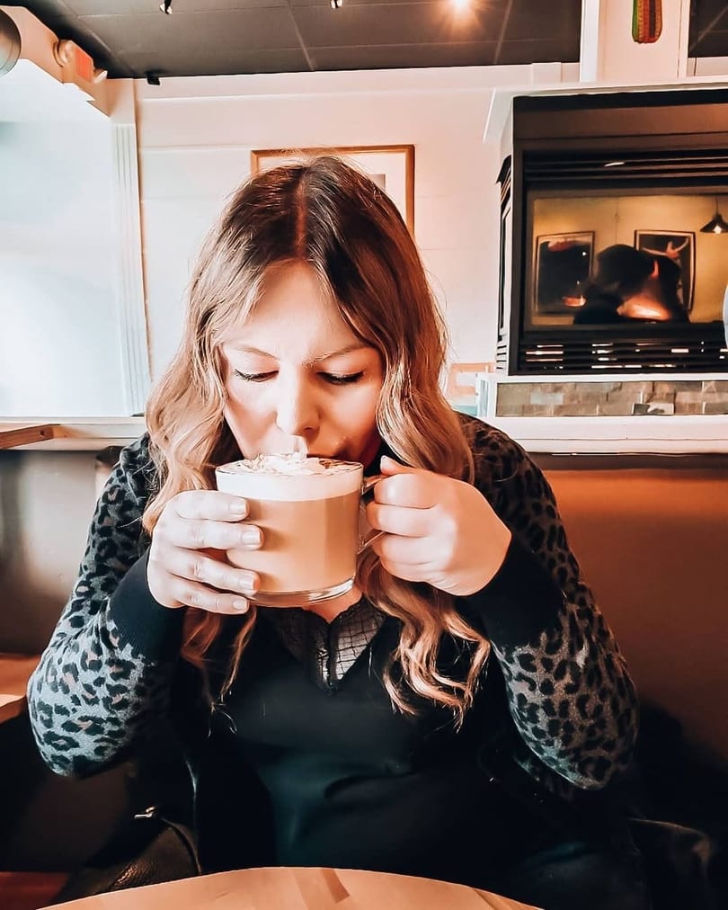 Woman in a leopard print sweater sips a frothy drink from a clear mug in a cozy restaurant.
