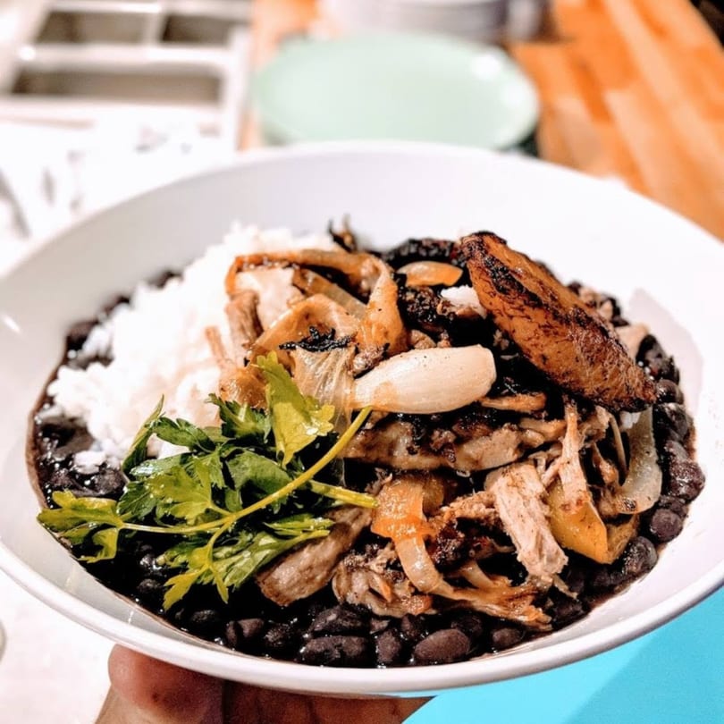 A white bowl filled with black beans, white rice, shredded meat, onions, and a plantain.